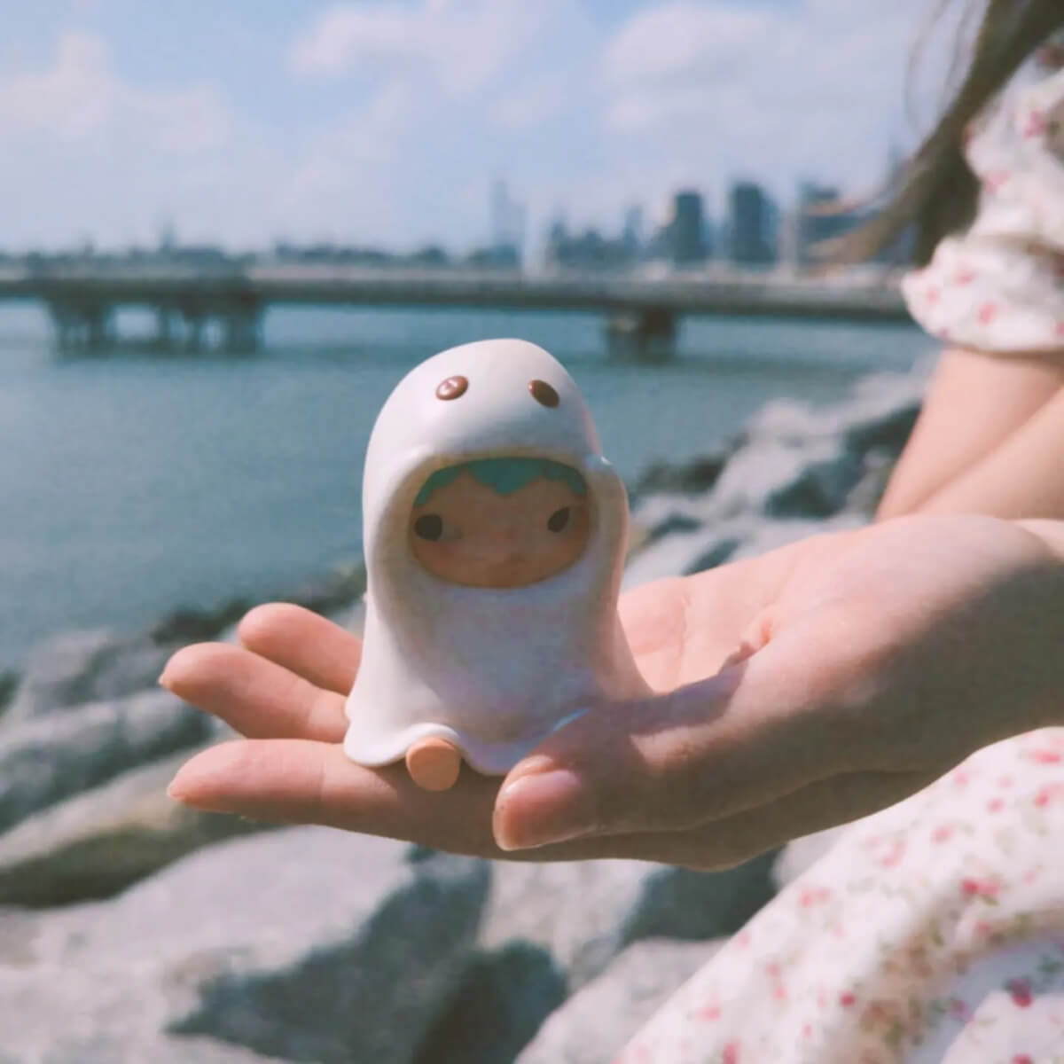 Woman sitting next to a river and holding a mini figure of a boy wearing a ghost costume